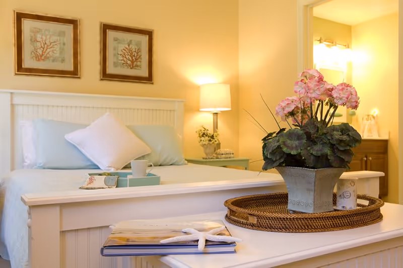 A cozy bedroom with a white wooden bed frame, light blue bedding, and a white pillow. On the bed is a tray with a cup and a small book. A bedside table with a lamp and a small plant is visible in the background. In the foreground, there is a white bench with a wicker tray holding a potted plant with pink flowers and a small container. Two framed coral prints hang on the wall above the bed. A bathroom with a sink and mirror is partially visible through an open door.