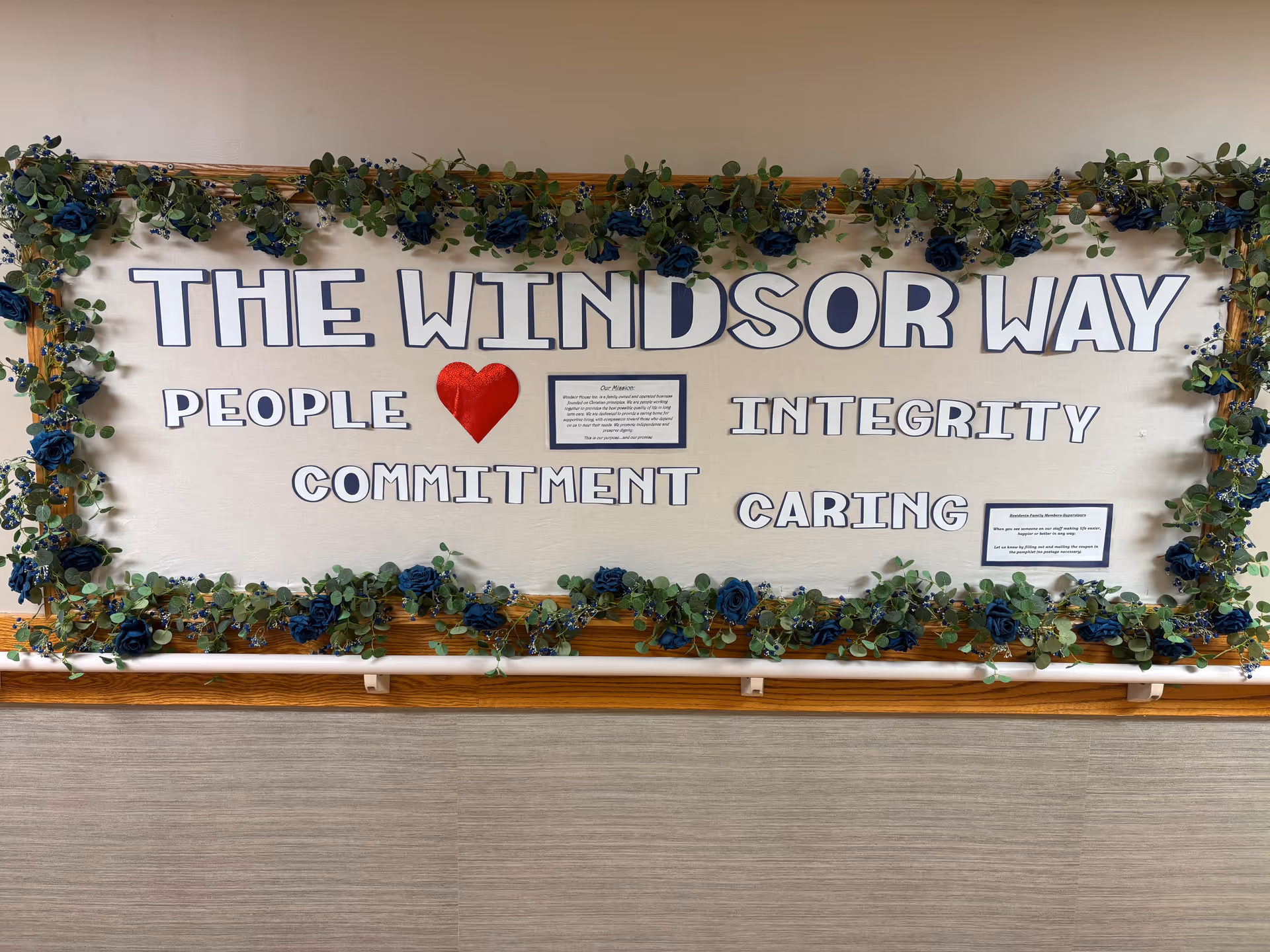 A decorative bulletin board with the title 'THE WINDSOR WAY' surrounded by green foliage and blue flowers. The board features the words PEOPLE, INTEGRITY, COMMITMENT, and CARING, with a red heart symbol between PEOPLE and INTEGRITY. There are two small framed texts on the board.