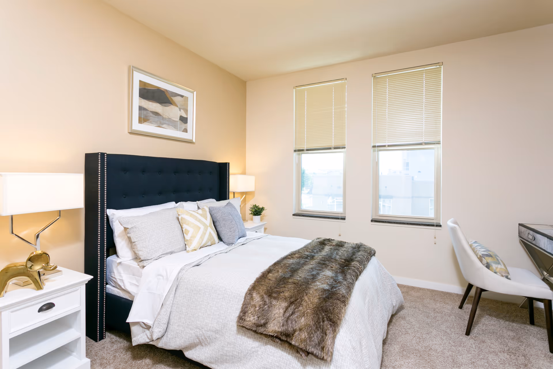A neatly made bed with a dark upholstered headboard, white and patterned pillows, and a faux fur throw blanket in a bright bedroom with beige walls, two windows with blinds, a white nightstand with a lamp and decorative golden elephant, and a white chair next to a desk.
