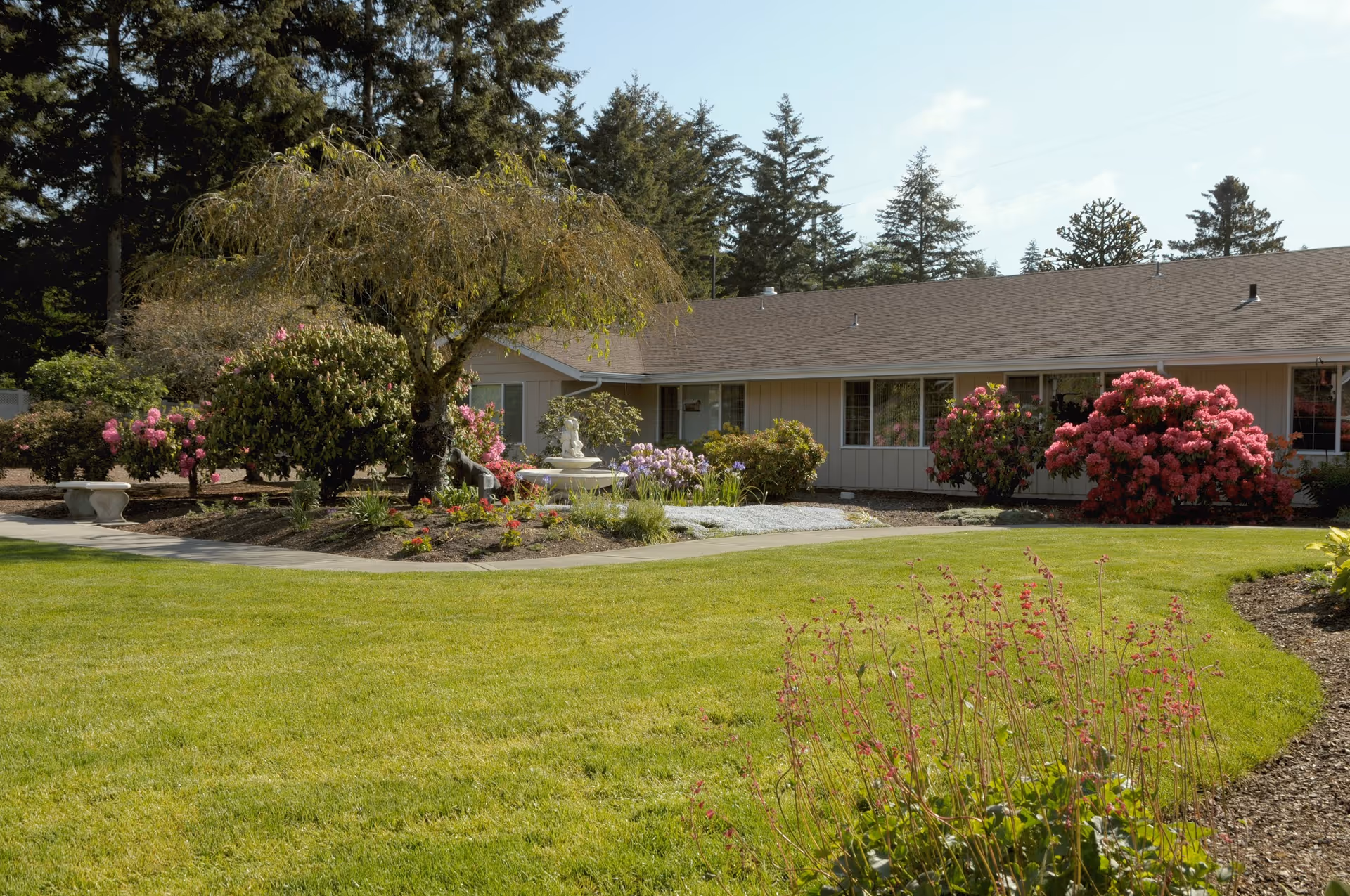 A well-maintained garden area with green grass, flowering bushes, and a small tree in front of a single-story building with beige siding and a brown roof. The garden includes a white stone bench and a white fountain surrounded by flowers. Tall evergreen trees are visible in the background under a partly cloudy sky.