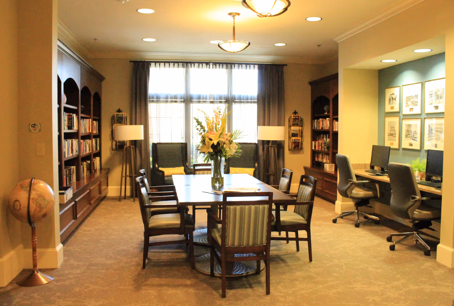 A well-lit common area with a large wooden table surrounded by chairs, a vase with flowers in the center, bookshelves filled with books on both sides, two armchairs near a window with curtains, and a small workspace with computers and office chairs in a recessed area on the right.