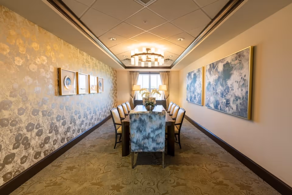 Well-lit formal dining room with a long table, upholstered chairs, decorative wallpaper and framed artwork.