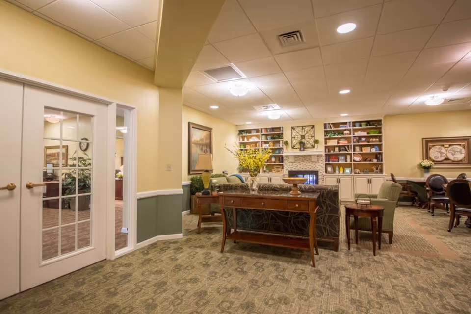 A cozy living room area in a senior living facility with a patterned carpet, a wooden console table with a vase of yellow flowers, a sofa, armchairs, and built-in bookshelves surrounding a fireplace. The walls are painted yellow with a green lower half, and there are framed artworks and a lamp on a side table. Double glass doors lead to another room.