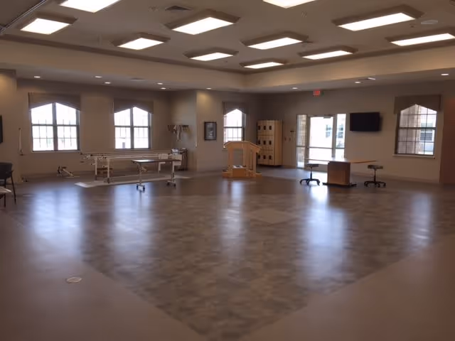 A spacious, well-lit room with large windows, a few chairs, a table, and some medical or rehabilitation equipment, likely used for physical therapy or group activities in a healthcare facility.
