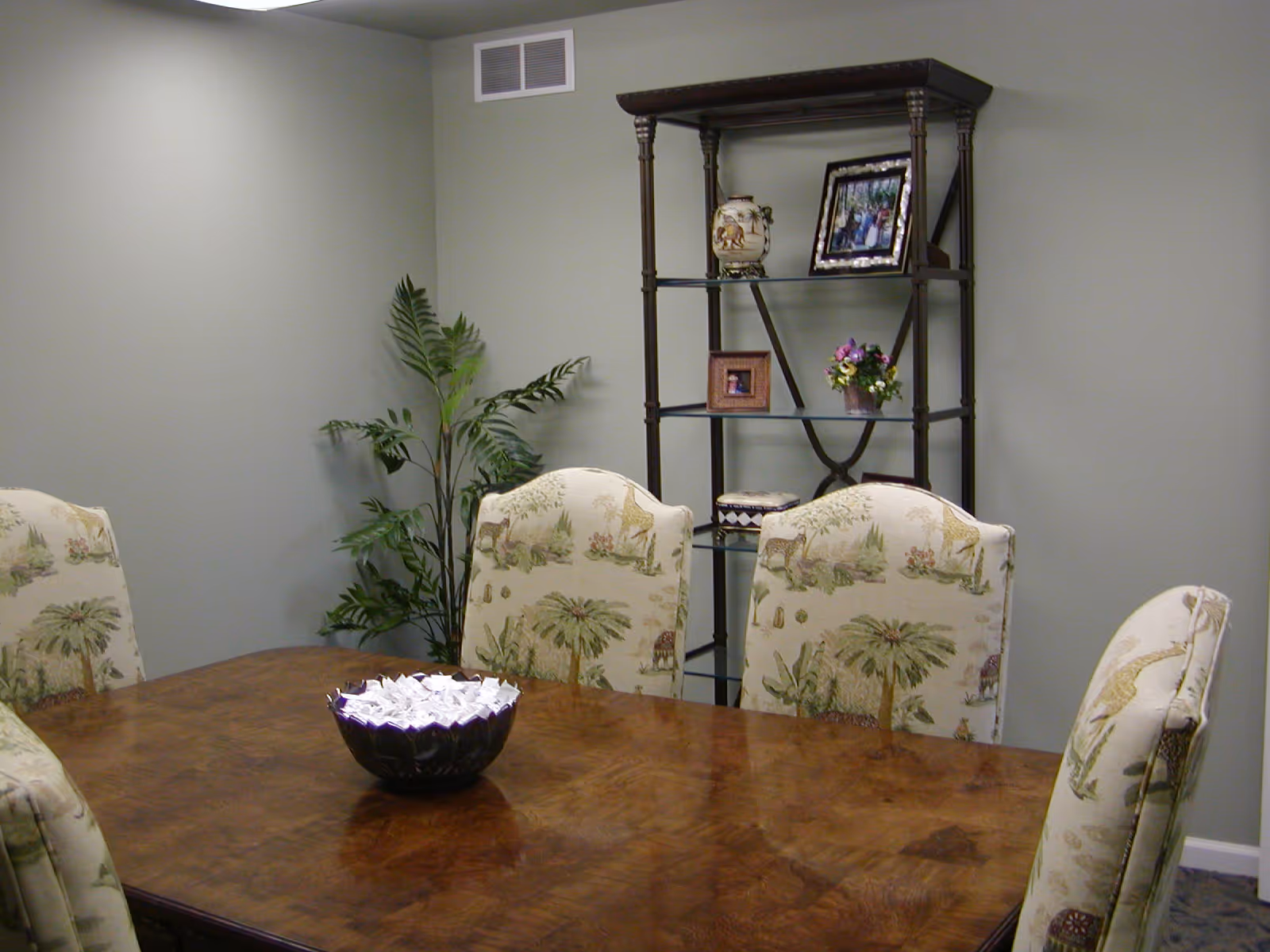 A dining room with a wooden table surrounded by six upholstered chairs featuring a nature-themed pattern. A decorative bowl filled with white paper cranes sits on the table. In the background, there is a black metal shelving unit with framed photos, a vase, and a small flower arrangement. A green leafy plant is placed in the corner against a light gray wall.