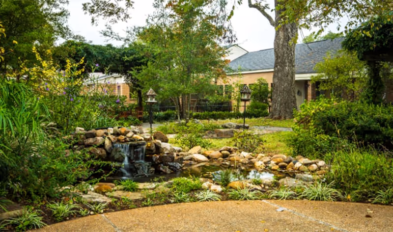 Landscaped outdoor courtyard with a small rock waterfall and pond surrounded by greenery and a brick building in the background.