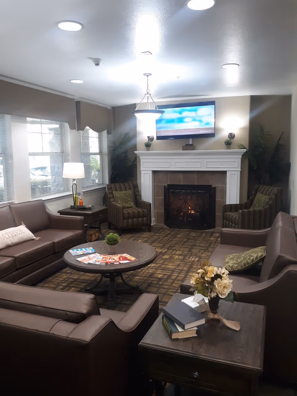A cozy living room with brown leather sofas and striped armchairs arranged around a round wooden coffee table. The room features a fireplace with a fire burning and a flat-screen TV mounted above it. There are windows with blinds on the left side, a table lamp on a side table, and a small plant and magazines on the coffee table. A side table in the foreground holds a vase with flowers and some books.