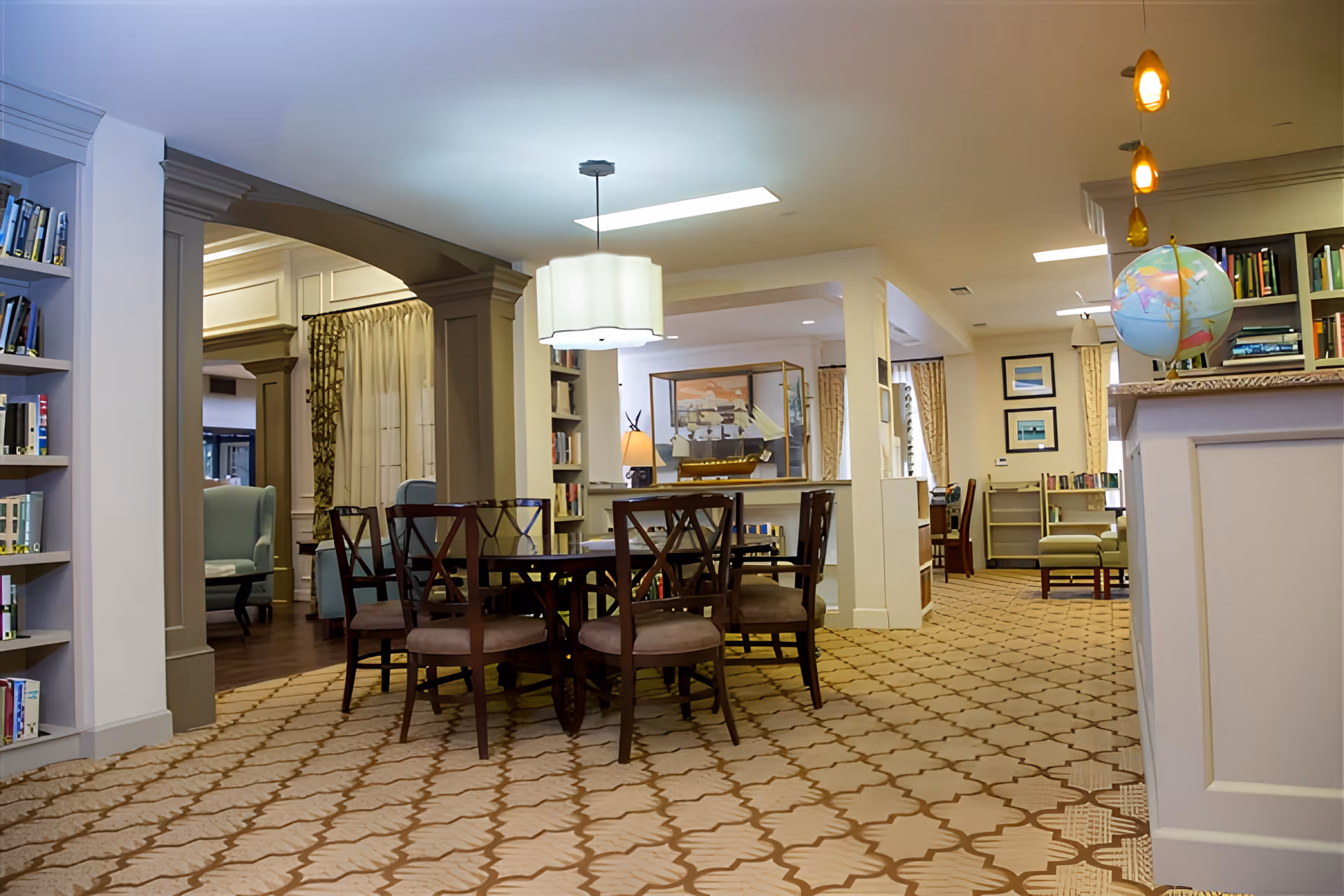 Interior view of a senior living facility common area with a round wooden table surrounded by six chairs on a patterned carpet. Bookshelves filled with books line the walls, and there are armchairs and framed pictures in the background. A globe sits on a counter to the right under hanging pendant lights.