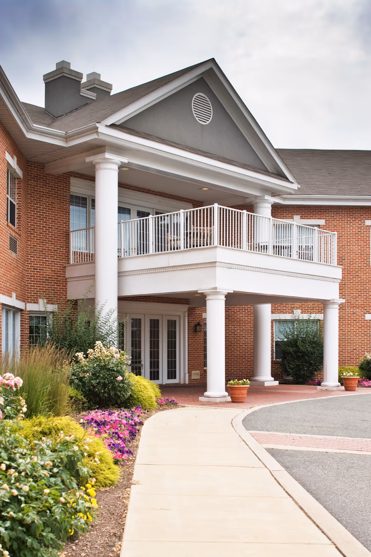 Entrance of a brick building with white columns supporting a balcony above. There is a curved sidewalk leading to double glass doors, surrounded by landscaped bushes and colorful flowers.