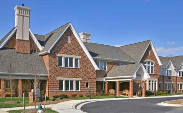 Exterior view of a large brick senior living facility building with multiple peaked roofs, chimneys, and several windows. The entrance has a covered porch with seating, and there is a curved driveway in front. The sky is clear and blue.