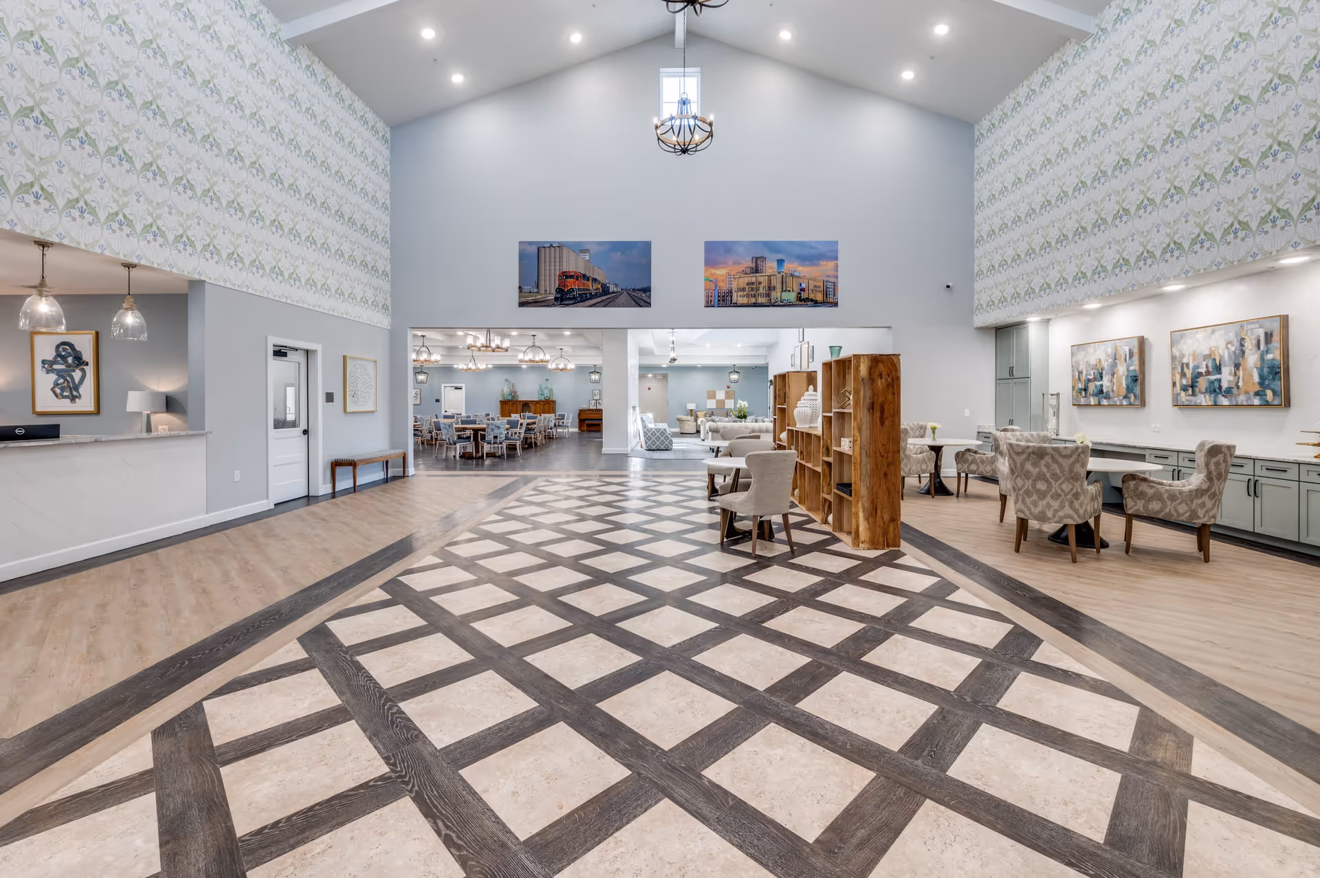 Spacious interior common area of The Lodge of Saginaw Health & Wellness featuring a high vaulted ceiling with a chandelier, patterned flooring, seating areas with armchairs and tables, decorative wall art, and a reception desk on the left side.