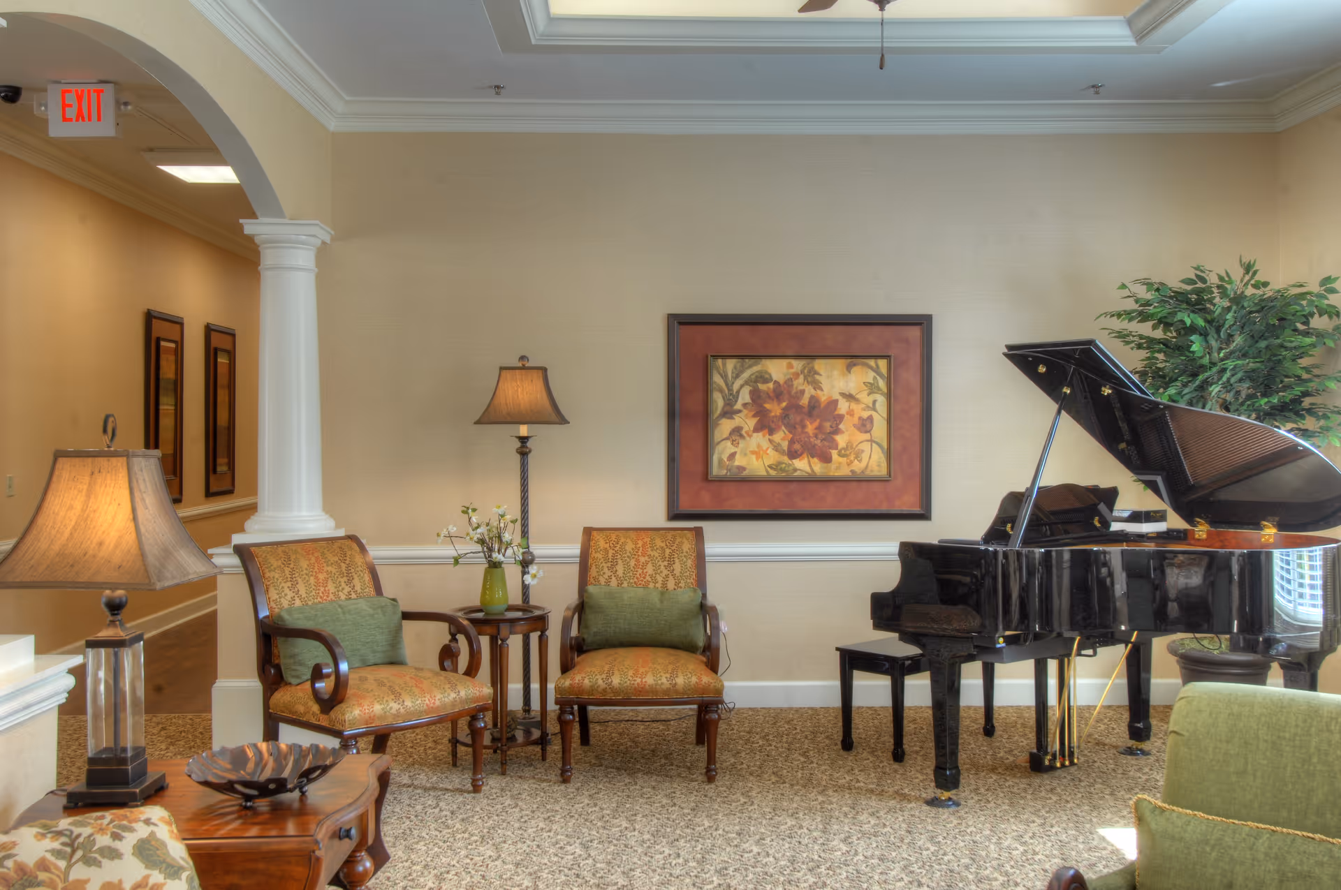 A cozy common room with a grand piano, upholstered chairs, side tables, lamps, and framed artwork on the wall.