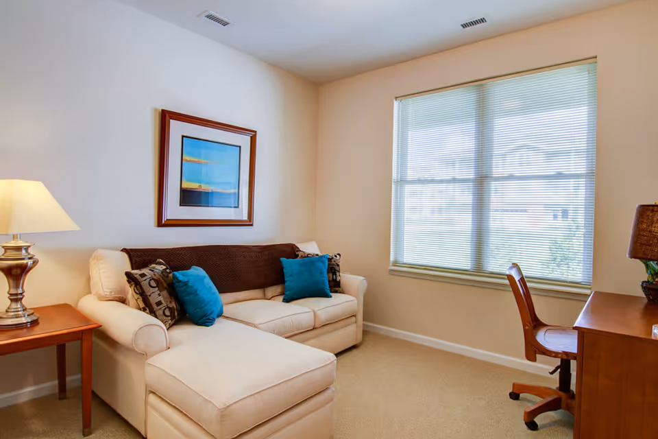 Light-filled living room with a beige chaise sofa accented by blue pillows, a side table lamp, framed artwork, a desk and a large window with blinds.