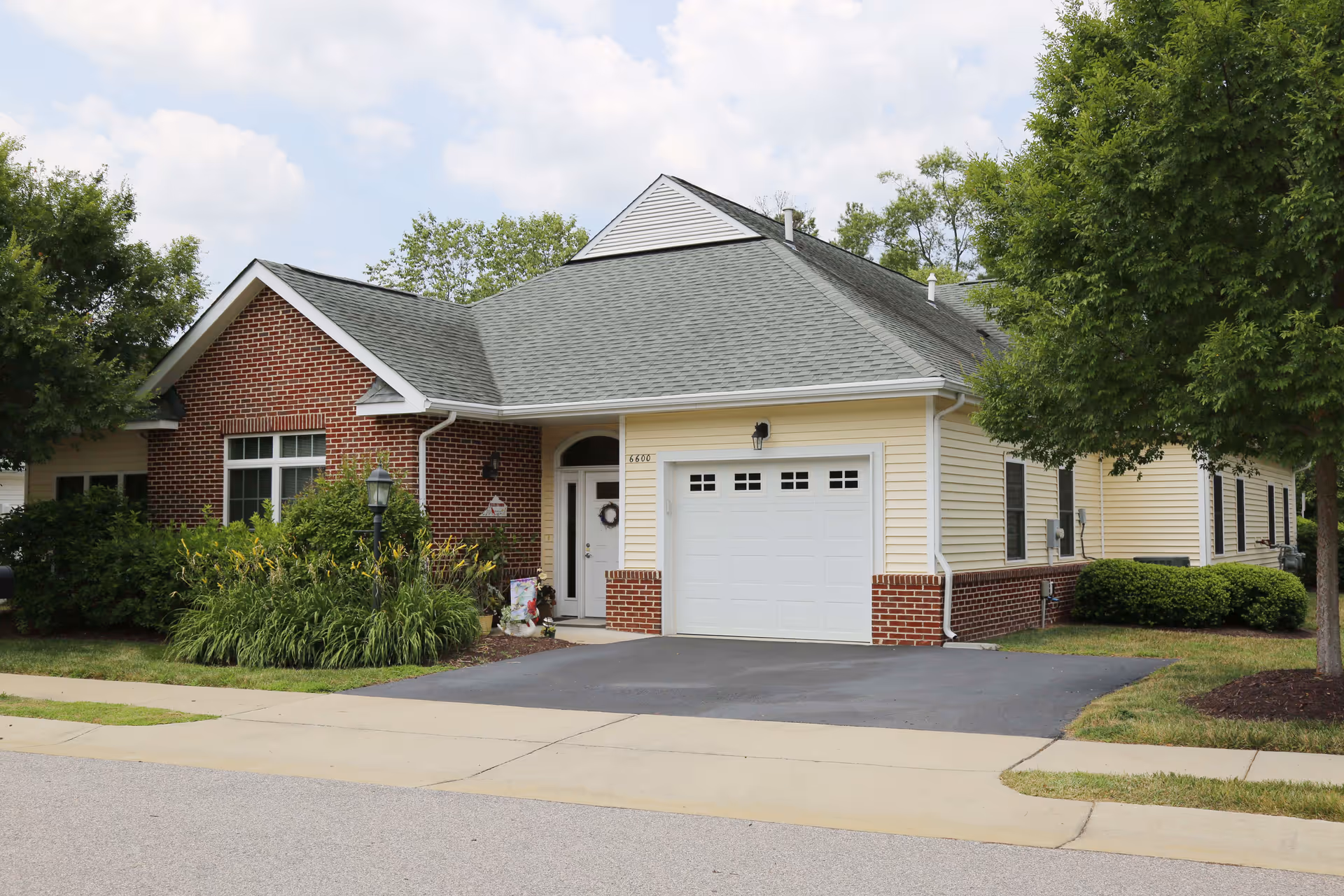 Exterior view of a single-story residential building with a combination of red brick and light yellow siding. The building features a gray shingled roof, a white garage door, a front door with a small wreath, and surrounding greenery including bushes and trees. The driveway and sidewalk are visible in the foreground under a partly cloudy sky.