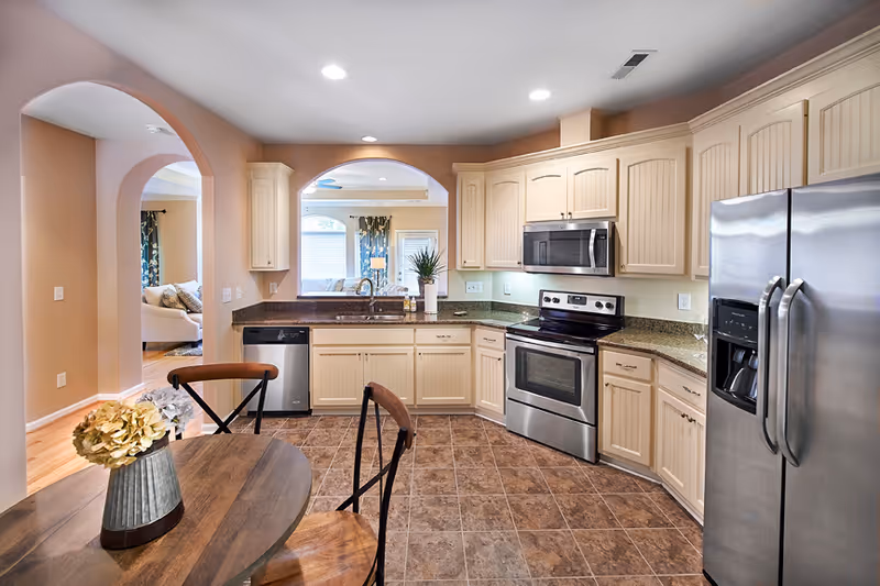 A modern kitchen with beige cabinets, granite countertops, stainless steel appliances including a refrigerator, oven, microwave, and dishwasher. There is a round wooden table with two chairs and a vase with flowers on it. The kitchen opens into a living room area visible through an arched doorway.