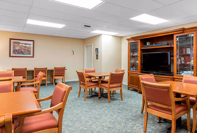 A common area with several wooden tables and chairs with red upholstery arranged on a blue patterned carpet. There is a large wooden entertainment center with a flat-screen TV and shelves containing books and decorative items. The walls are beige with a framed picture and a small cross hanging. The ceiling has recessed lighting.