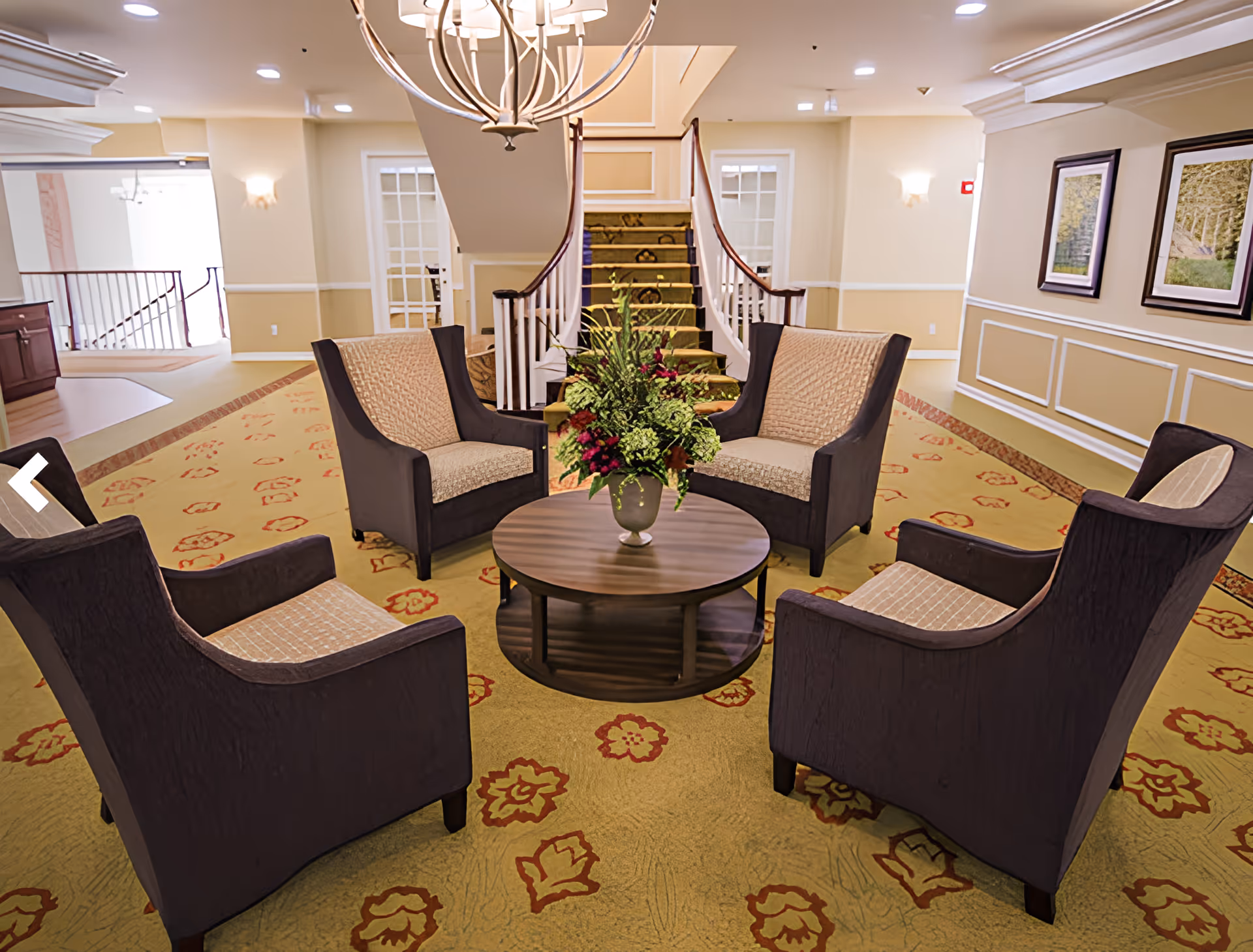 Bright lobby lounge with four armchairs arranged around a round coffee table and a central staircase in the background.