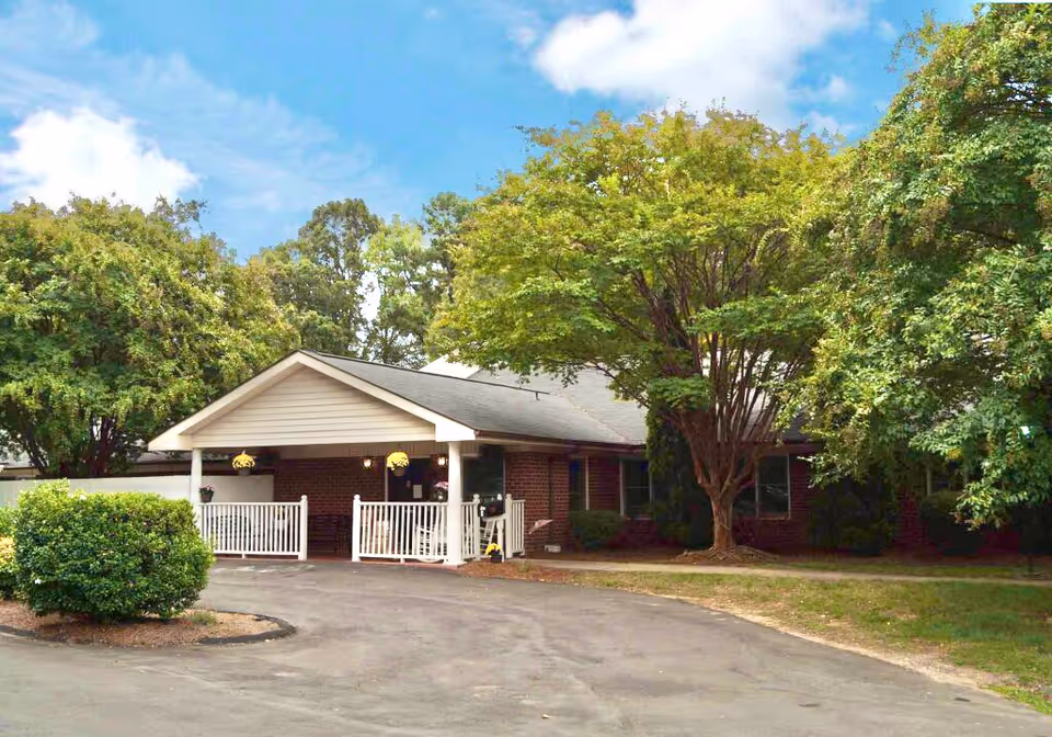 Single-story brick building with a covered white-railed entrance, driveway, and large trees surrounding it.