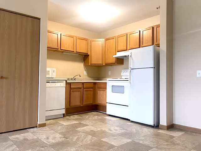 A small kitchen area with wooden cabinets, a white refrigerator, a white stove with an oven, a white dishwasher, and a white microwave. The floor is tiled with beige and light brown tiles, and there is a wooden door on the left side of the image.