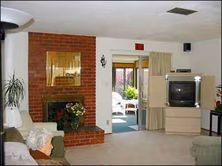 Elderly resident sits in Sunset Home for the Elderly living room with brick fireplace and TV.