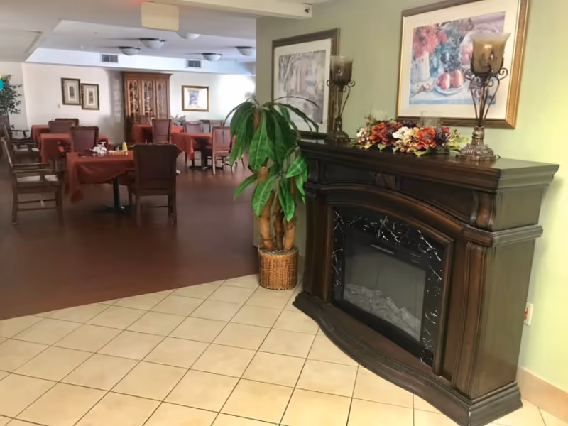 Interior view of a senior living facility dining area with tables covered in red tablecloths and wooden chairs. In the foreground, there is a dark wooden electric fireplace with decorative items on top, including two candle holders and a floral arrangement. A potted plant is placed next to the fireplace, and framed artwork hangs on the walls.