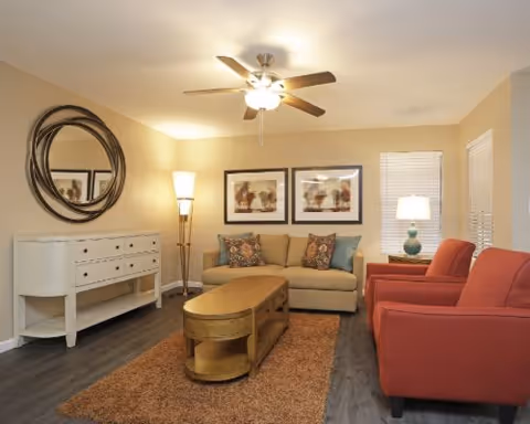 A cozy living room with a beige sofa adorned with decorative pillows, two red armchairs, a wooden coffee table on a brown rug, a white sideboard with drawers, a round decorative mirror above it, two framed artworks on the wall, a floor lamp, a table lamp on a side table, and a ceiling fan with lights.