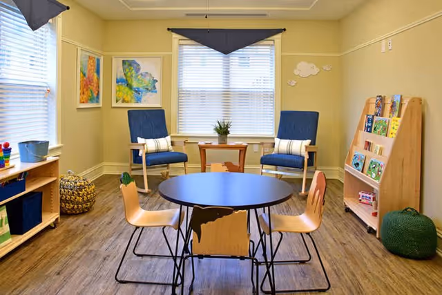 Bright activity/reading room with a round table and child-sized chairs, two armchairs by a window, and a small bookshelf of books.