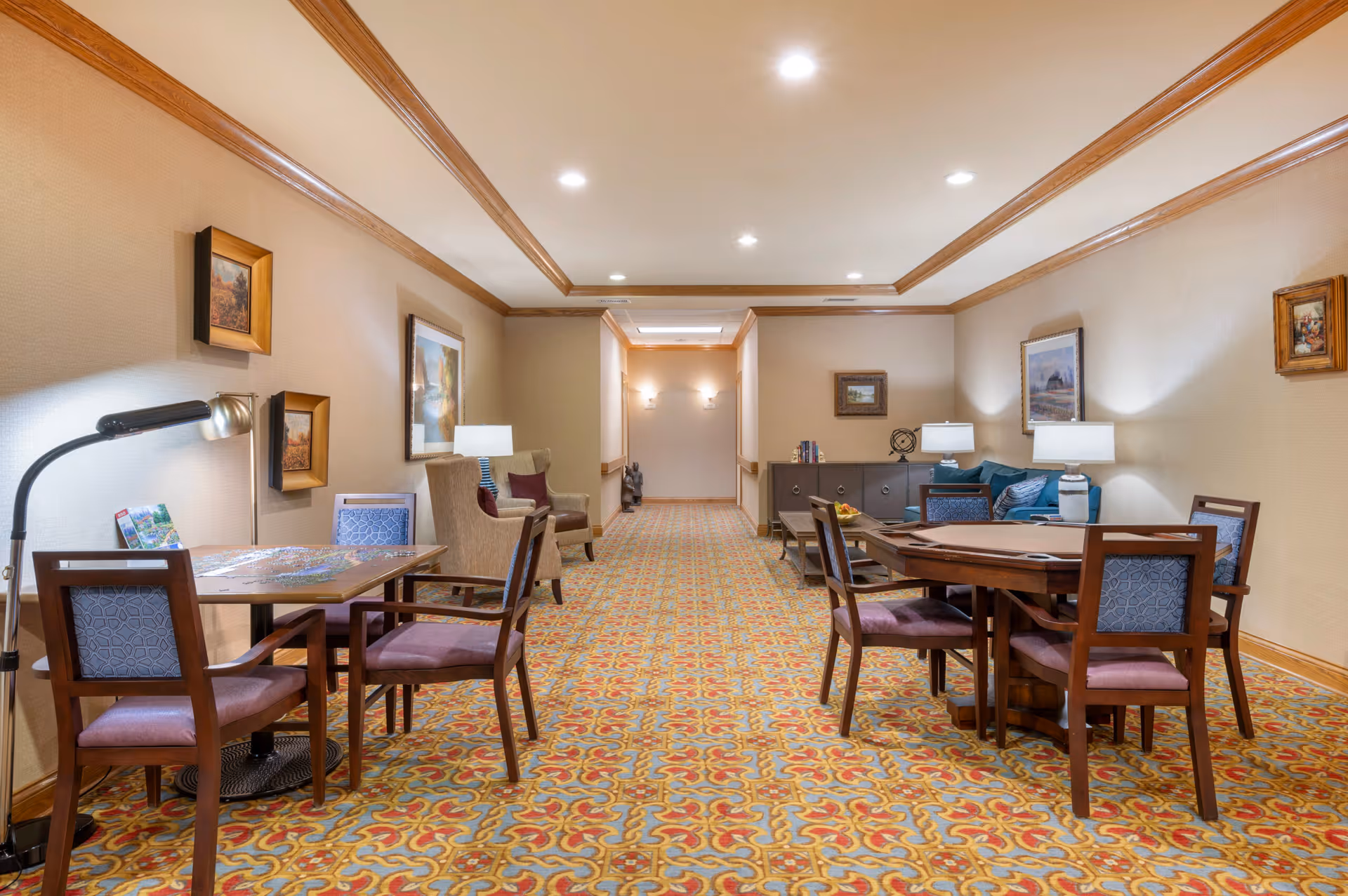 Well-lit communal lounge with multiple tables and chairs, armchairs, lamps, and framed artwork along a carpeted corridor.