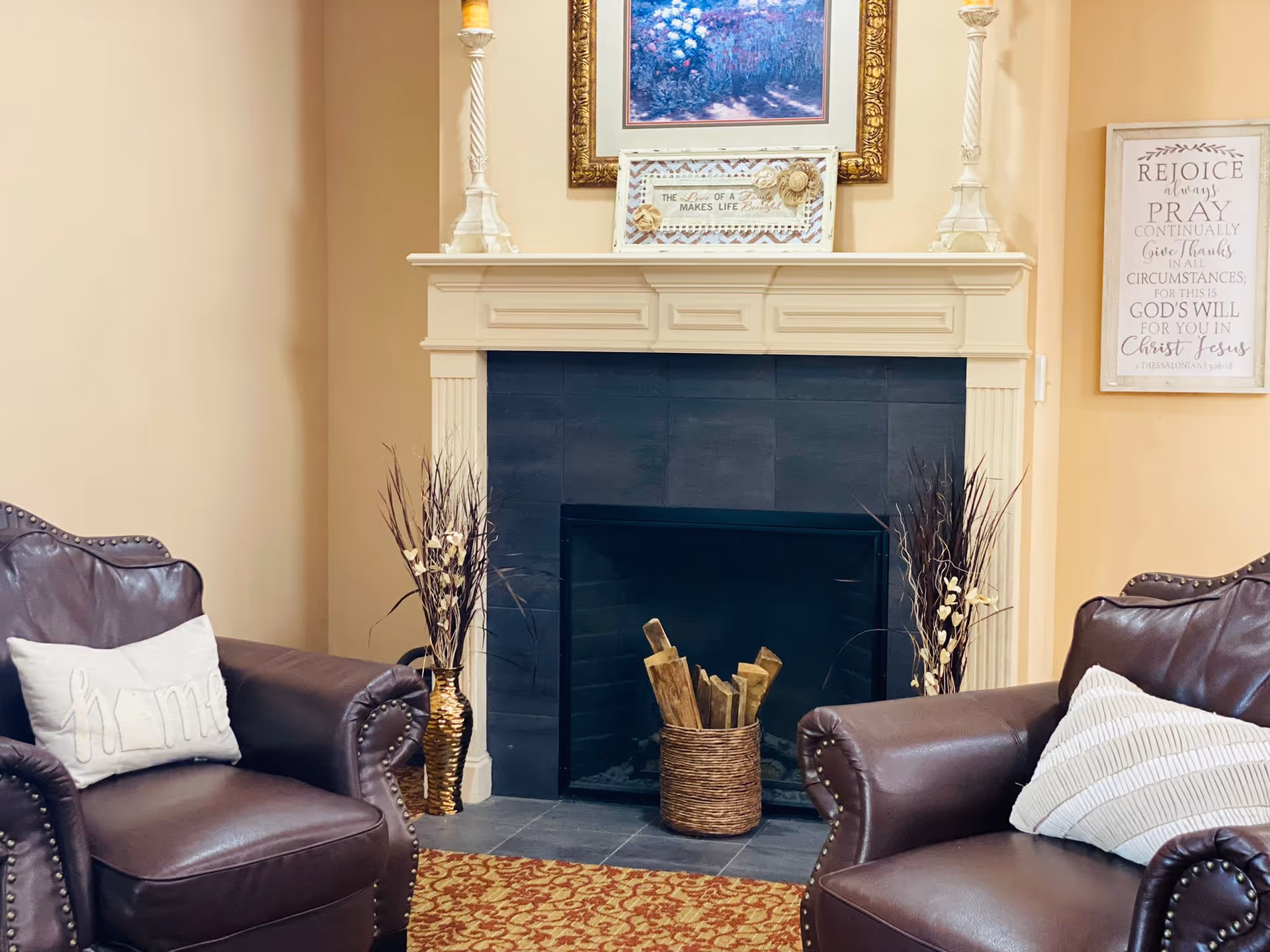 Cozy living room area with two brown leather armchairs, each with a decorative pillow. Between the chairs is a fireplace with a white mantel, black tile surround, and a basket of firewood inside. On the mantel are two tall candle holders, a framed picture, and a decorative sign. On the wall to the right is a framed inspirational quote. The floor has a patterned rug in front of the fireplace.