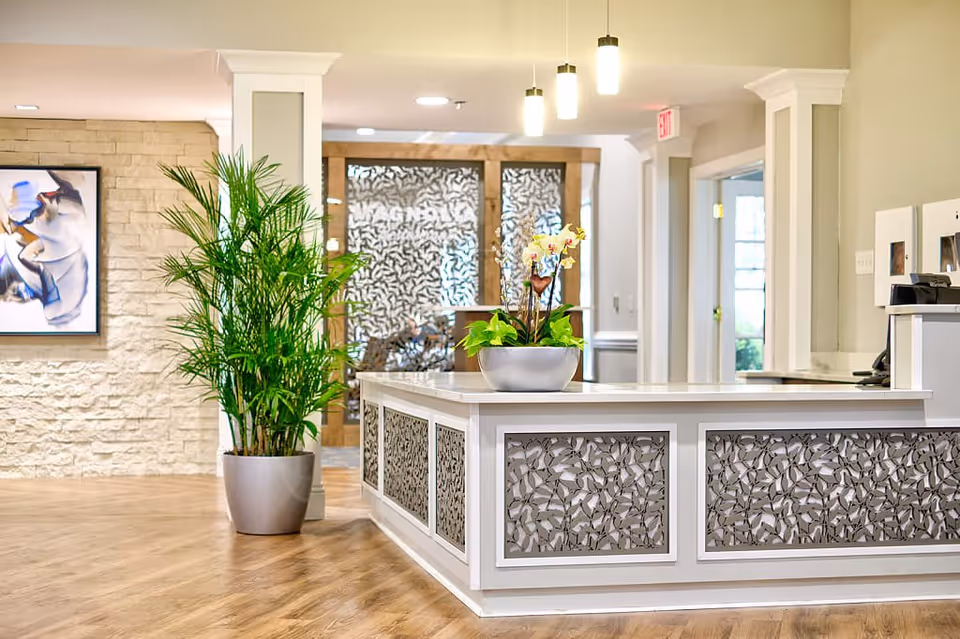 Bright and modern reception area with a white decorative front desk featuring intricate leaf patterns, a potted green plant on the floor to the left, and a flower arrangement on the desk. The background includes a textured stone wall with abstract artwork and glass doors with frosted leaf designs.