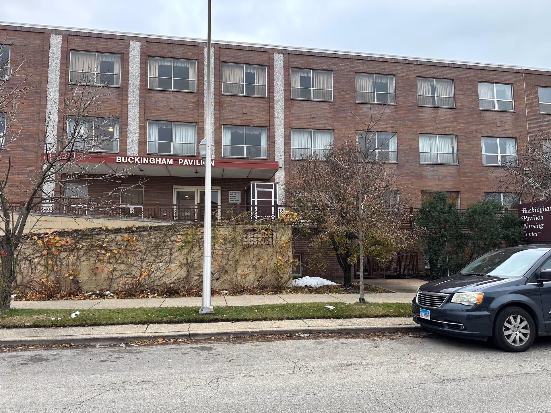 Exterior view of Buckingham Pavilion Nursing and Rehabilitation building, a three-story brick structure with multiple windows. There is a maroon awning above the entrance displaying the facility name. A black minivan is parked on the street in front of the building, and leafless trees and some greenery are visible along the sidewalk.