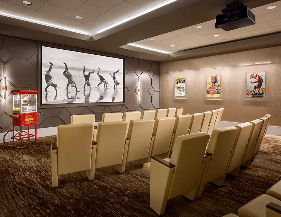 A cozy senior living facility movie theater room with beige cushioned seats arranged in rows facing a large screen displaying a black and white image of people doing handstands on a beach. The room has a popcorn machine on the left side and three vintage movie posters on the right wall. The lighting is warm and recessed in the ceiling.