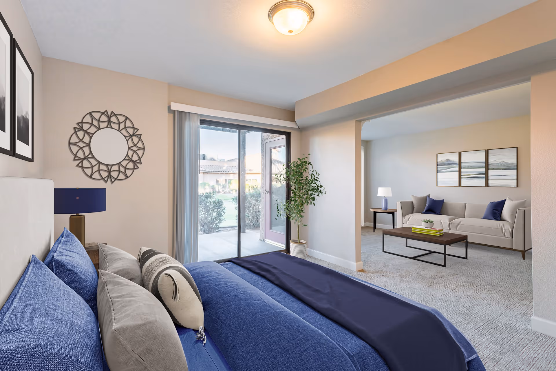 A bright senior living bedroom with a bed dressed in blue and gray bedding, a decorative round mirror on the wall, and a sliding glass door leading to an outdoor patio. Adjacent to the bedroom is a living room area with a beige sofa, a wooden coffee table, side tables with lamps, and framed artwork on the wall.