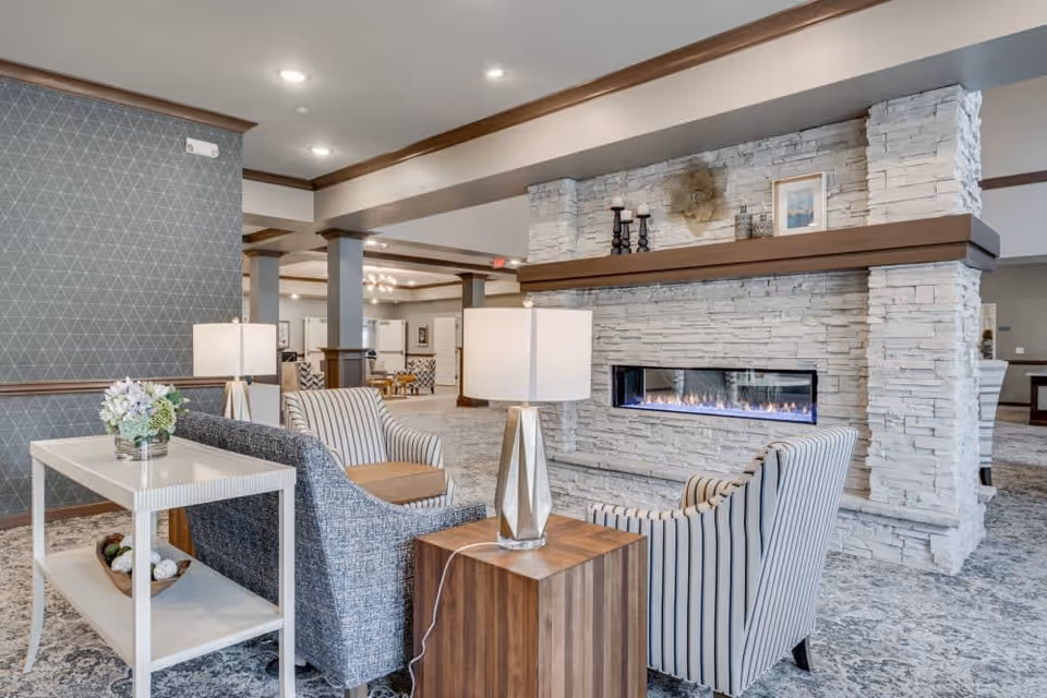 A cozy seating area in a senior living facility featuring two striped armchairs and one blue patterned armchair around a modern rectangular fireplace set in a white stone wall. There are two table lamps on wooden and white side tables, a floral arrangement on one table, and a patterned carpet covering the floor. The space has recessed lighting and decorative wall paneling.