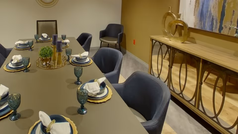 A formal dining room with a long table set with plates, napkins and teal glasses, surrounded by blue upholstered chairs and a decorative sideboard.