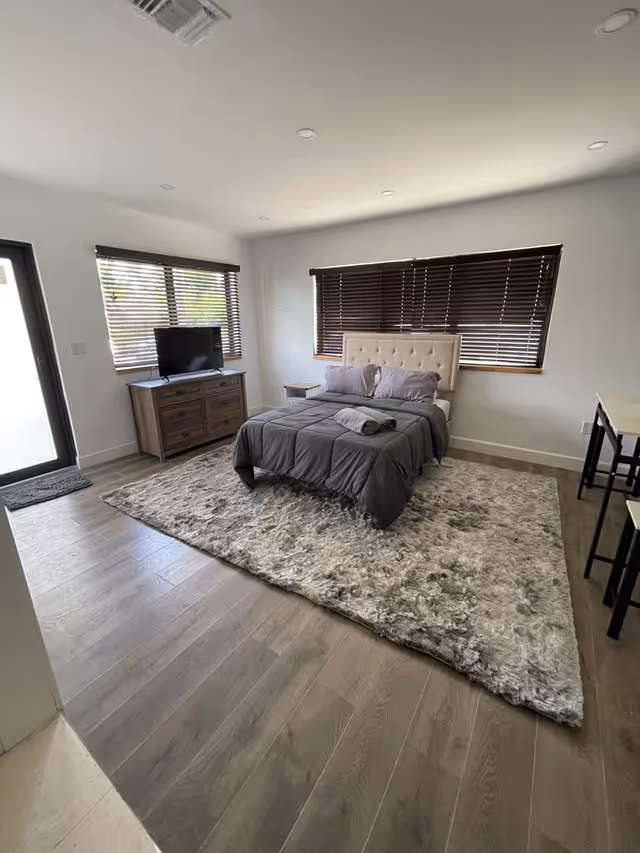 A bright bedroom with a large bed covered in gray bedding and two pillows, placed on a fluffy gray area rug. There is a wooden dresser with a flat-screen TV on top, two windows with dark blinds, a small nightstand next to the bed, and a door with a window on the left side. The room has light-colored walls and wooden flooring.