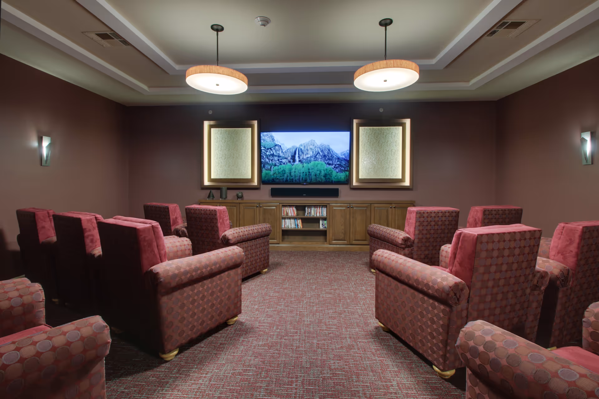 A cozy senior living facility media room with multiple upholstered armchairs arranged in rows facing a large flat-screen TV mounted on a wall. The room has warm brown walls, two ceiling lights, and a wooden cabinet below the TV holding books and decorative items.