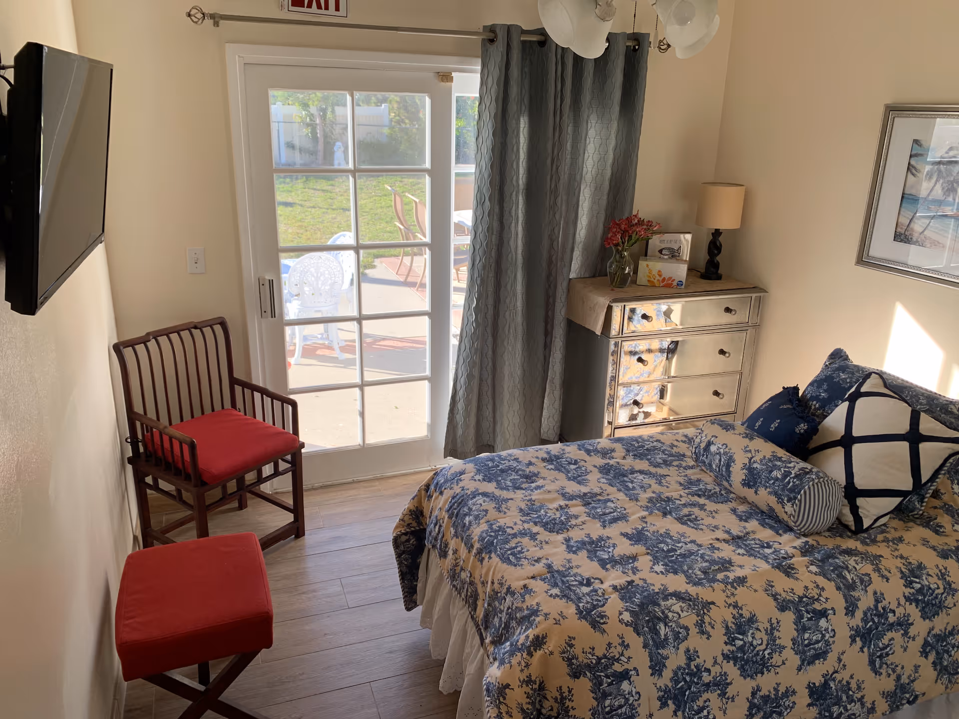 A cozy bedroom with a bed covered in a blue and beige patterned bedspread and several pillows. There is a mirrored dresser with a lamp and flowers on top, a wooden chair with a red cushion, and a matching red cushioned stool. A flat-screen TV is mounted on the wall. The room has a glass door with curtains leading to an outdoor patio area with chairs and a grassy yard.