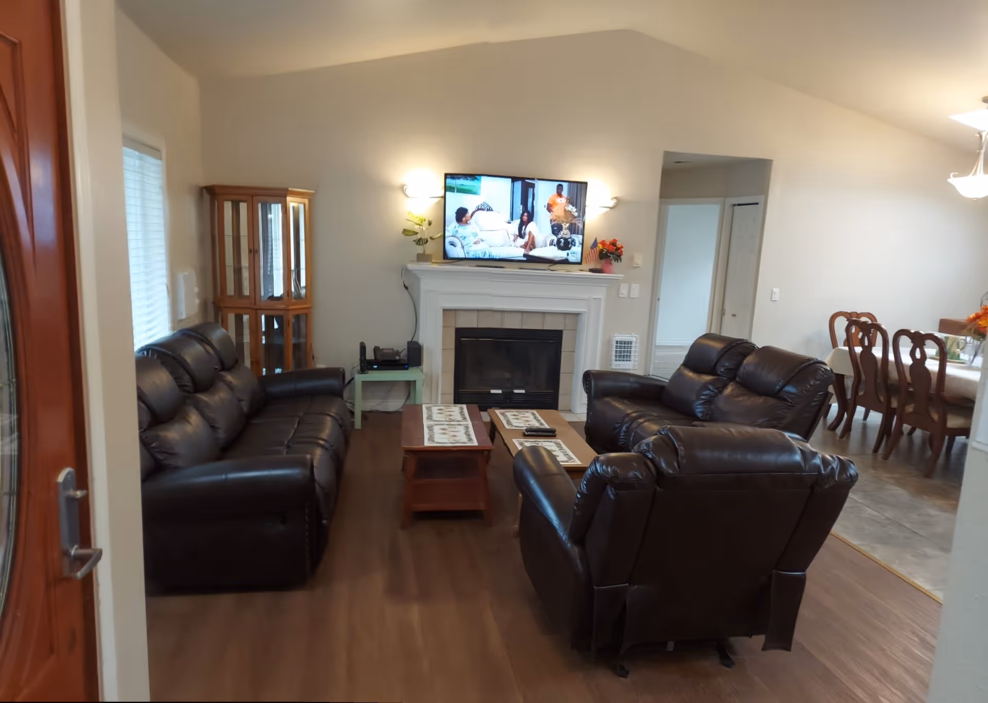 Interior view of a living room with dark brown leather sofas arranged around a wooden coffee table. A flat-screen TV is mounted above a white fireplace, with a small green side table and a wooden display cabinet to the left. The room has wooden flooring and is adjacent to a dining area with a table and chairs.