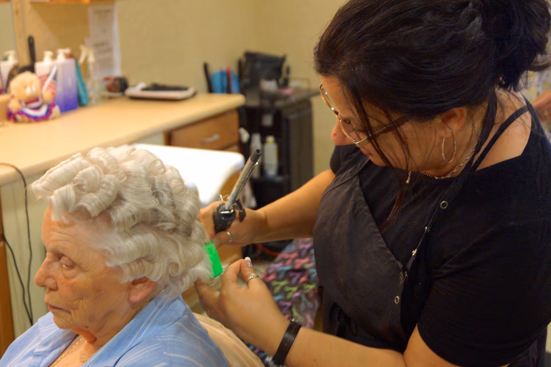 An elderly woman with white hair is having her hair styled with a curling iron by a hairstylist in a salon setting. The stylist is focused on curling the woman's hair, and various salon tools and products are visible in the background.