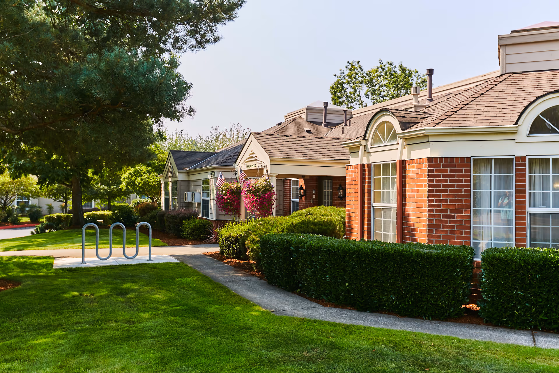 Exterior view of a senior living facility building with brick and beige siding, surrounded by well-maintained green lawns, bushes, and trees. There is a concrete pathway leading to the entrance, which is decorated with hanging flower baskets and American flags. A metal bike rack is visible on the lawn near the pathway.
