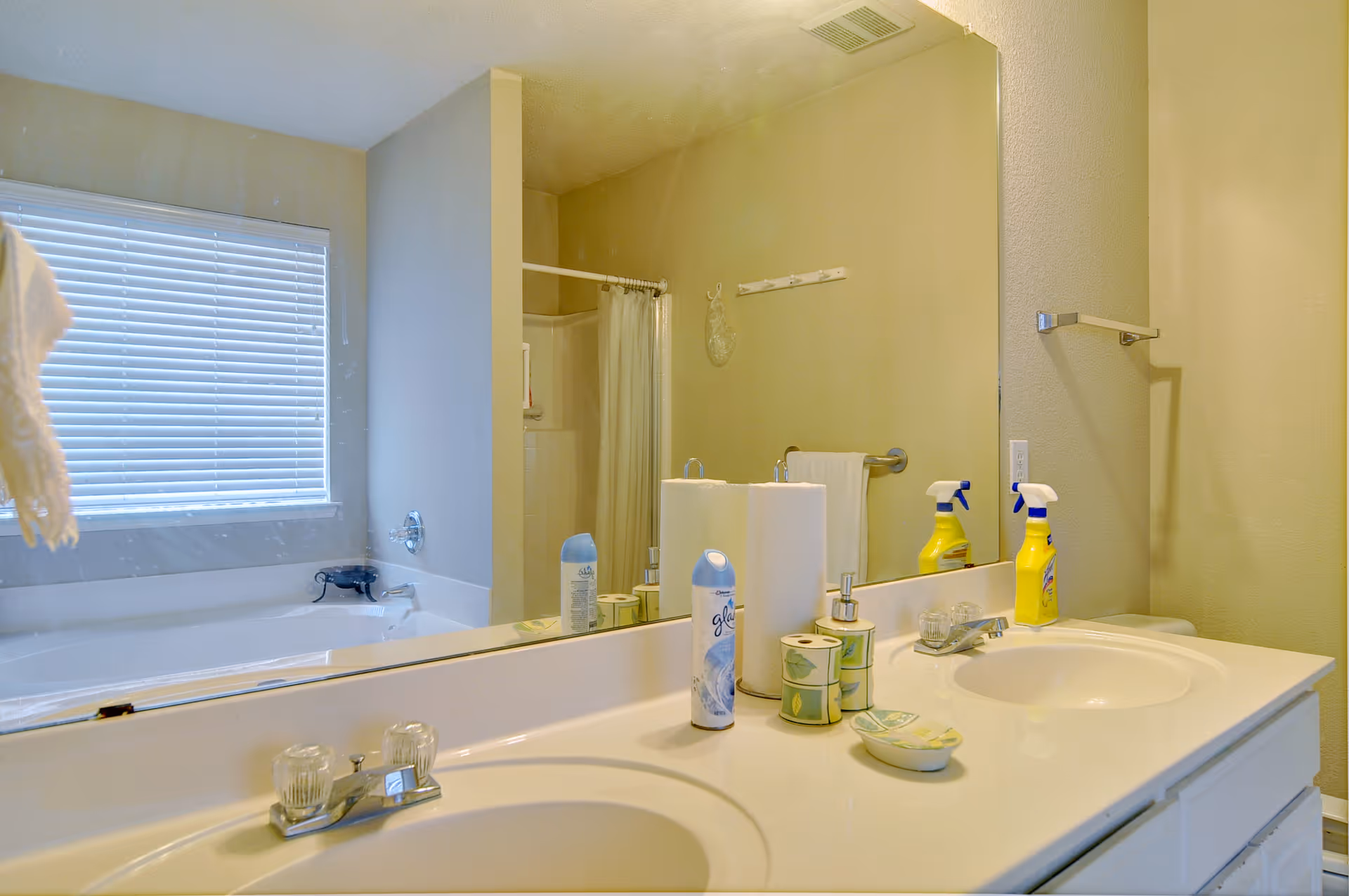 A bathroom with a double sink vanity, a large mirror, and various toiletries including air freshener, paper towels, soap dispenser, and cleaning spray. In the background, there is a bathtub with a window above it and a shower area with a curtain.