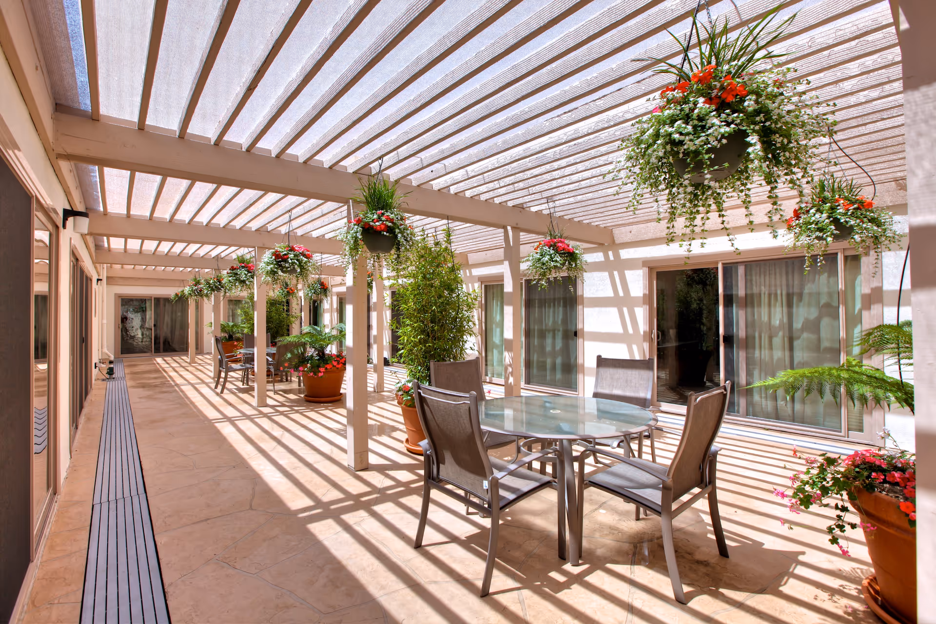 Outdoor patio area with a glass-top round table and four chairs under a pergola with hanging flower pots and potted plants along the sides. The patio is adjacent to a building with sliding glass doors and windows.