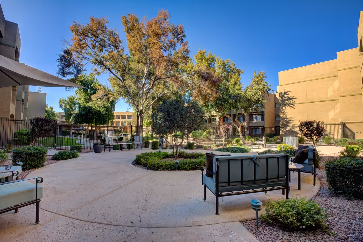 Outdoor courtyard area at Thunderbird Senior Living with patio seating, trees, shrubs, and a clear blue sky. The space is surrounded by buildings and has paved walkways and garden beds.