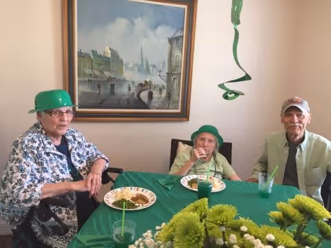 Three elderly individuals sitting at a table covered with a green tablecloth, wearing green hats. They have plates with food and drinks in front of them. A large framed painting of a cityscape hangs on the wall behind them. There is a green hanging decoration above the table and a bouquet of green and white flowers in the foreground.