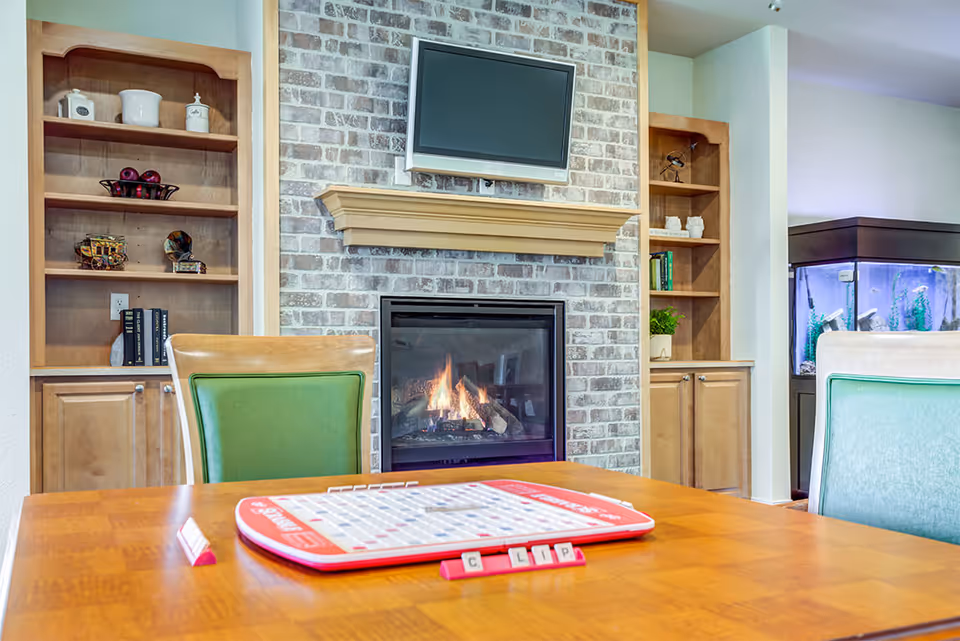 A cozy common room with a brick fireplace beneath a wall-mounted TV, flanked by wooden built-in shelves, an aquarium, and a table with a board game and green chairs.