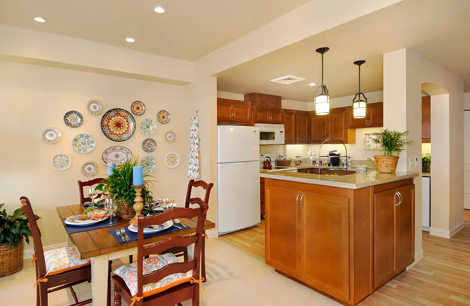 A cozy dining area with a wooden table set for four, decorated with blue candles and a green plant centerpiece. The wall behind the table features a decorative arrangement of colorful plates. Adjacent to the dining area is a kitchen with wooden cabinets, a white refrigerator, a microwave, and a kitchen island with a granite countertop and two hanging pendant lights.
