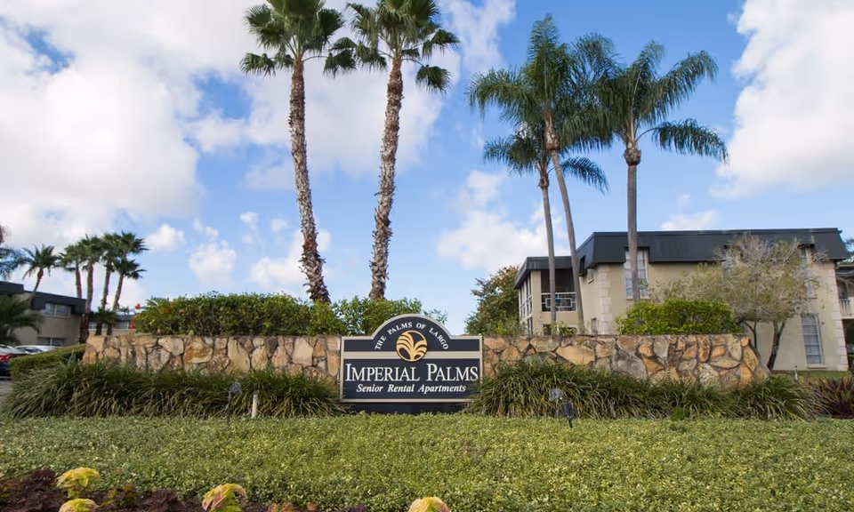Outdoor view of the entrance sign for Imperial Palms Senior Rental Apartments, surrounded by greenery and palm trees with apartment buildings in the background under a partly cloudy sky.
