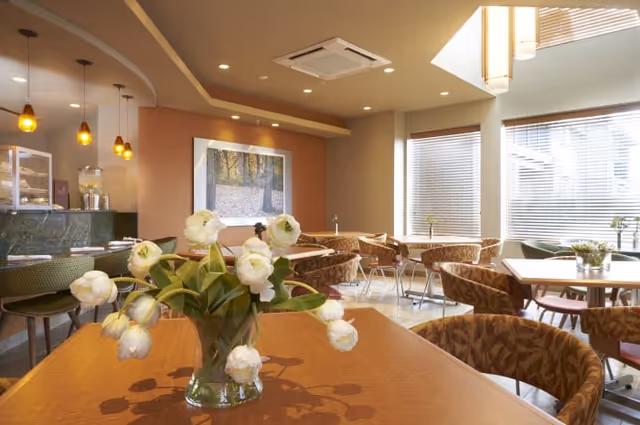 Dining area with a vase of white flowers on a foreground table, multiple round tables and chairs, pendant lights, and large windows.