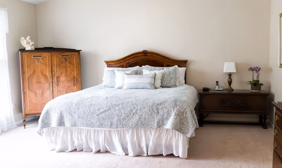 A neatly made bed with a wooden headboard, adorned with multiple pillows and a light blue patterned bedspread. To the left of the bed is a wooden cabinet with decorative items on top, and to the right is a wooden nightstand with a lamp, a phone, and a potted orchid. The room has light-colored walls and carpeted flooring.